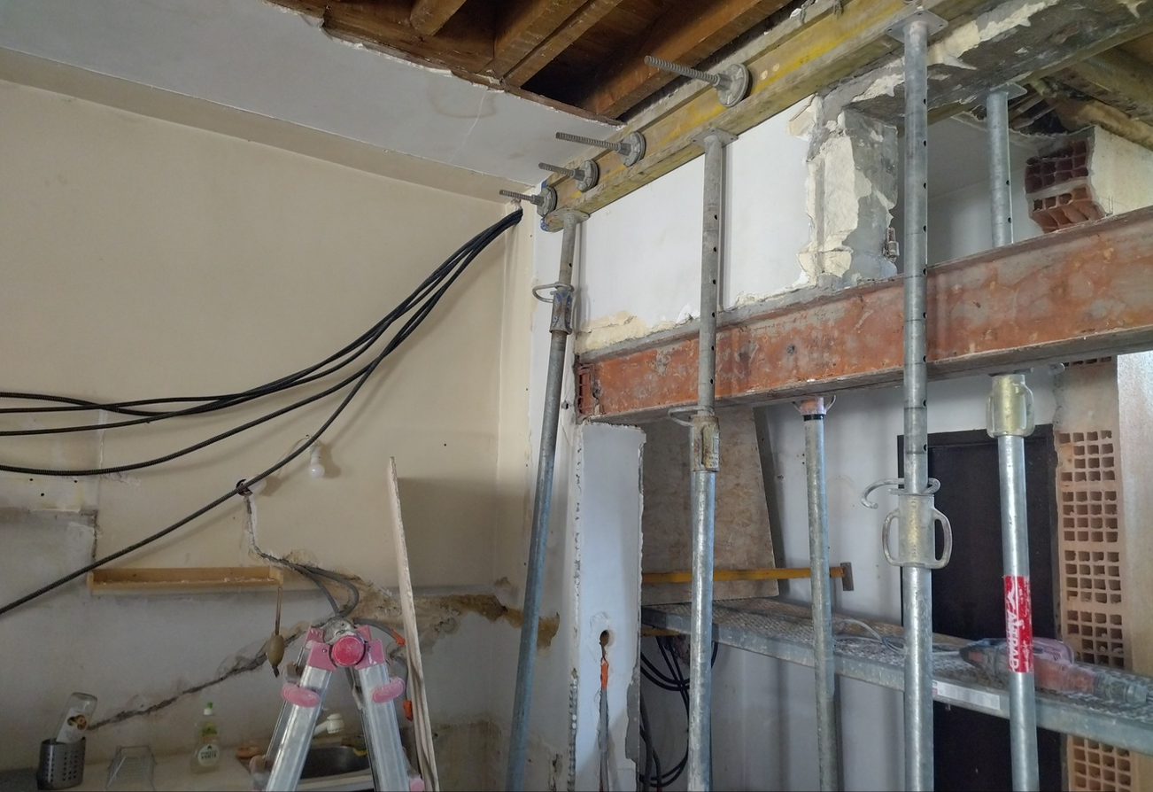 Chantier de rénovation avec poutre en acier rouillée soutenue par des étais métalliques, plafond exposé, câbles électriques et échelle.