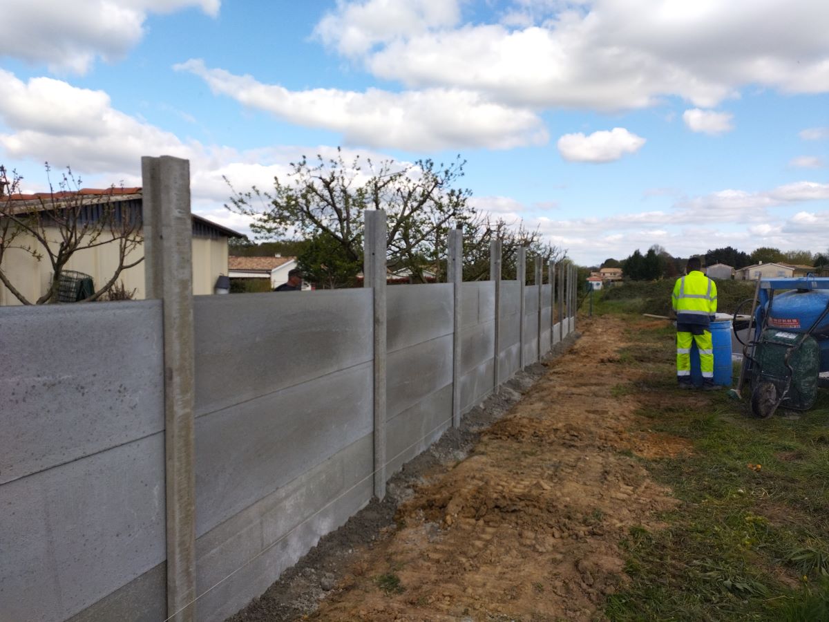 Installation d'une longue clôture en béton préfabriqué sur terrain nu. Un ouvrier en gilet jaune fluorescent observe le chantier.