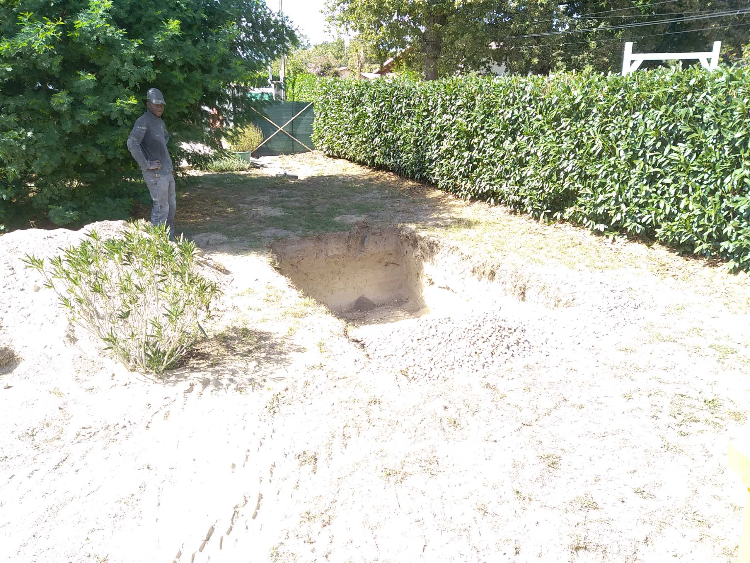 Ouvrier près d'un trou d'excavation et d'un tas de sable dans un jardin ensoleillé, avec haie verte et gravier.