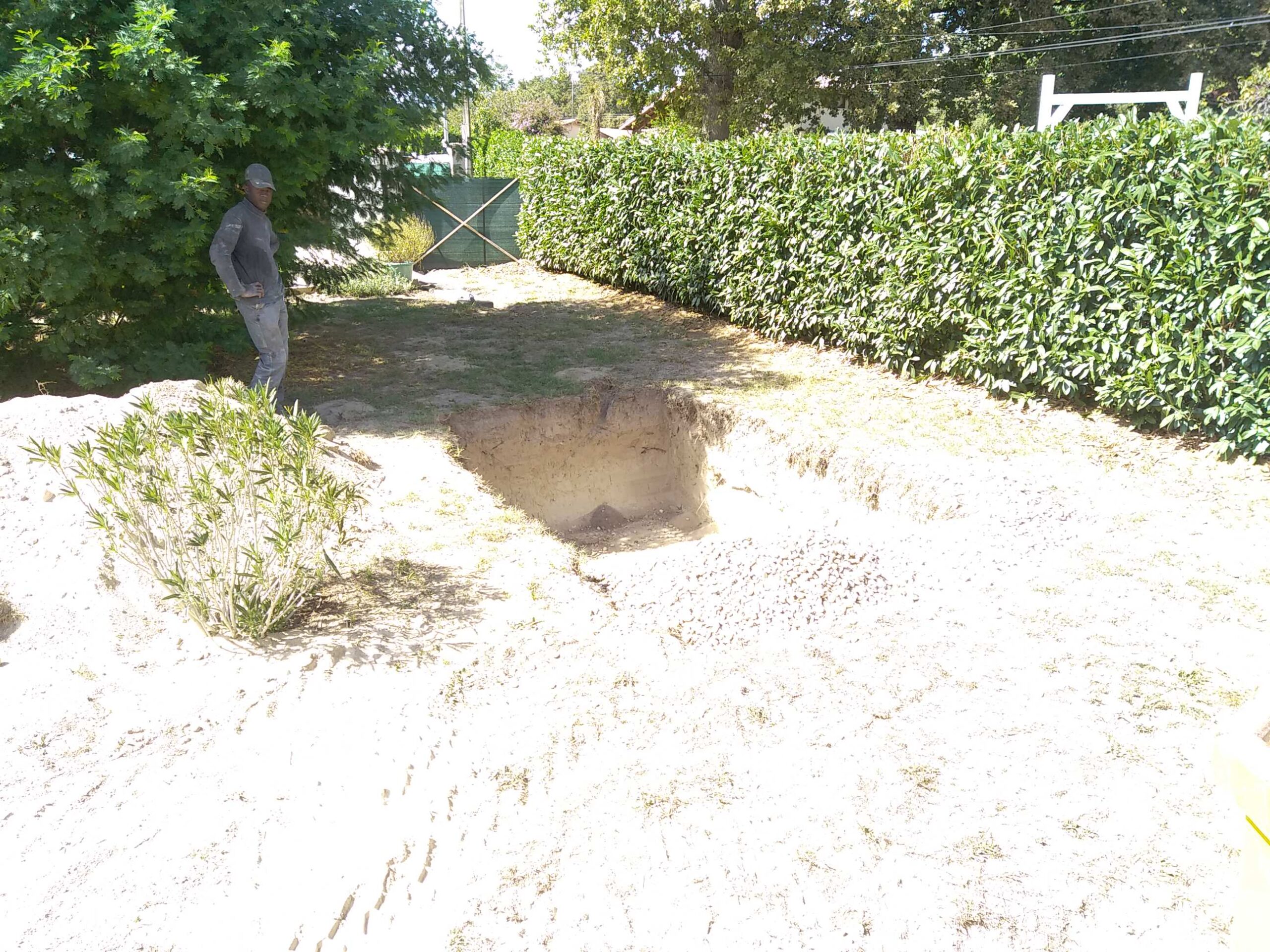 Homme inspectant une fosse rectangulaire creusée pour terrassement, avec tas de sable et verdure, dans un jardin ensoleillé.