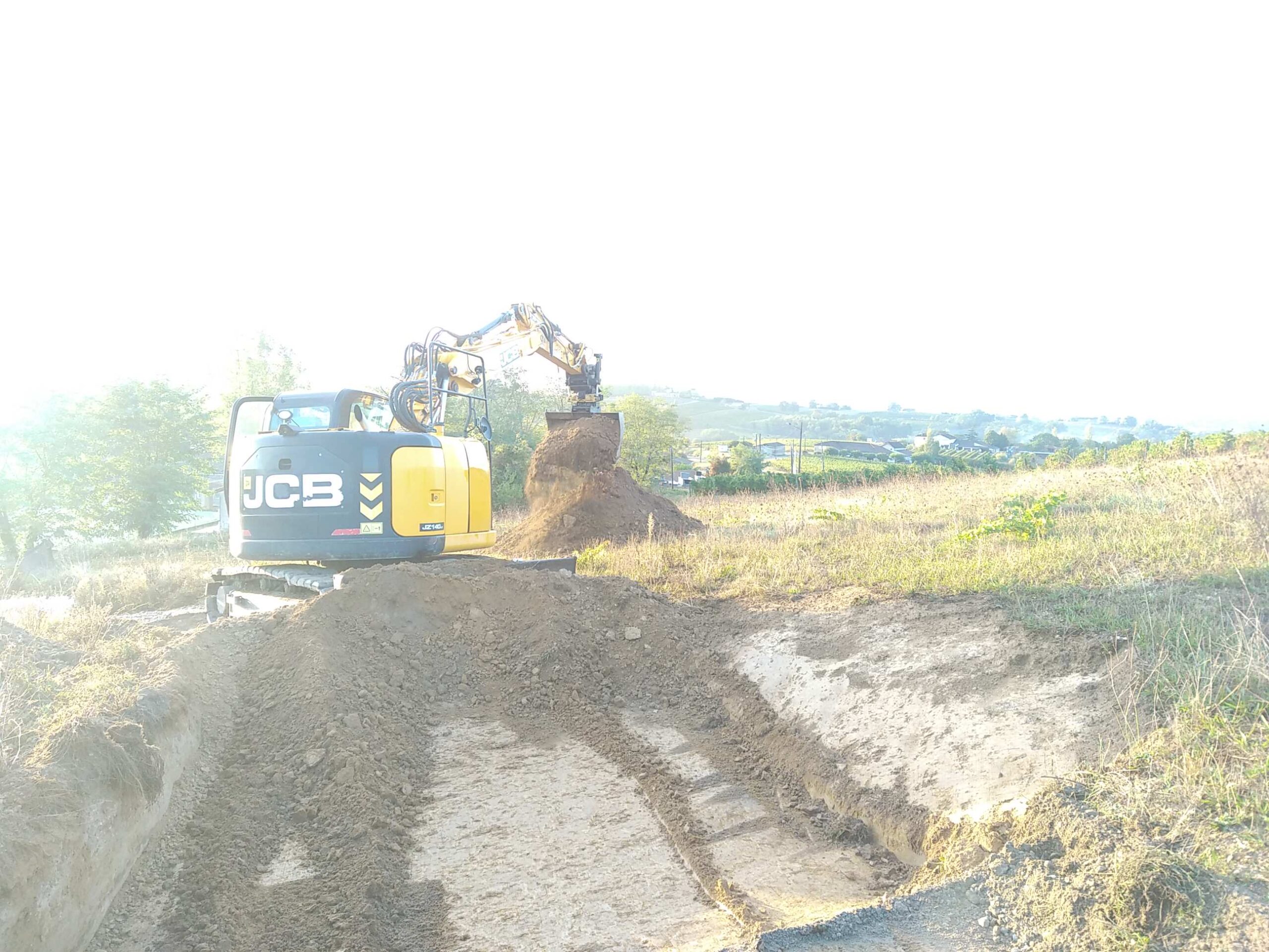 Pelle hydraulique JCB JZ140 jaune et noire creusant une tranchée dans un champ. La machine déplace de la terre, avec un tas de terre à côté.