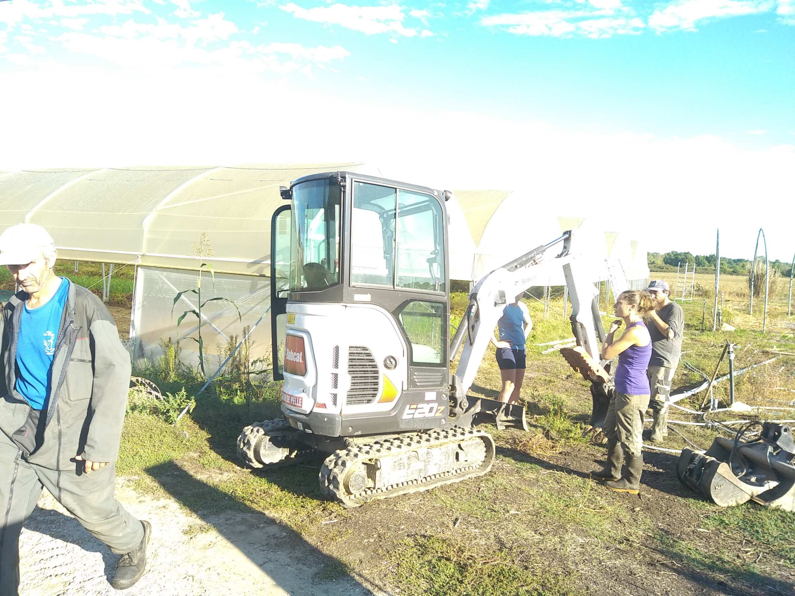 Mini-pelle Bobcat E20z sur terrain agricole avec serres. Des personnes discutent autour de l'engin blanc, un homme marche à gauche.