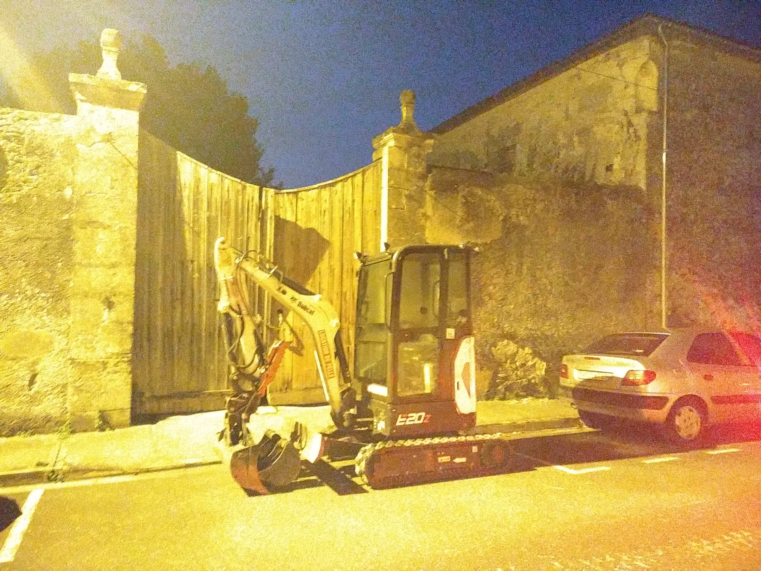 Mini-pelle Bobcat E20 jaune sur route au crépuscule. Mur pierre, grand portail bois courbé, voiture garée sous lumière urbaine.