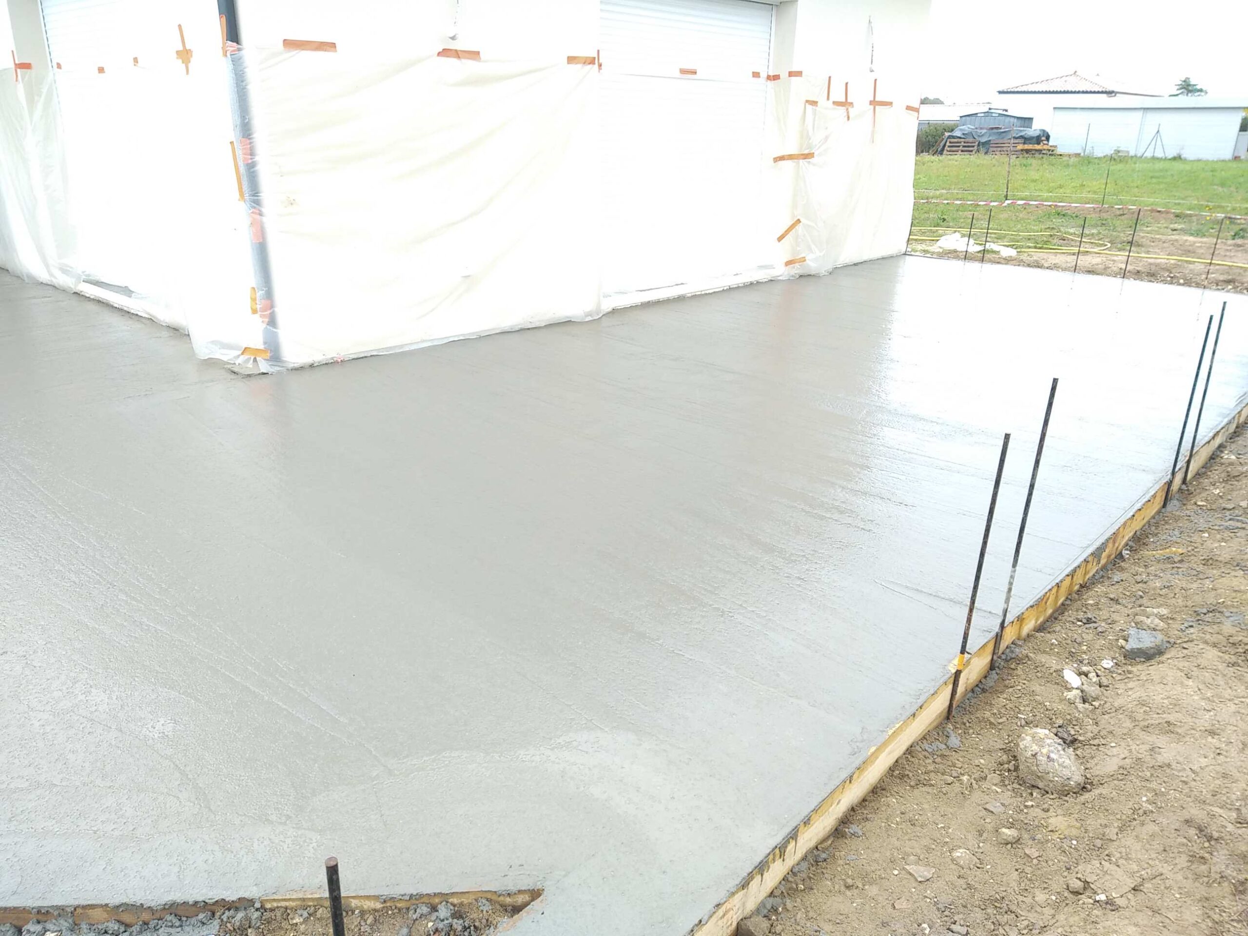 Dalle de béton grise fraîchement coulée, brillante et lisse, devant un bâtiment aux murs bâchés. Piquets métalliques bordent le chantier.