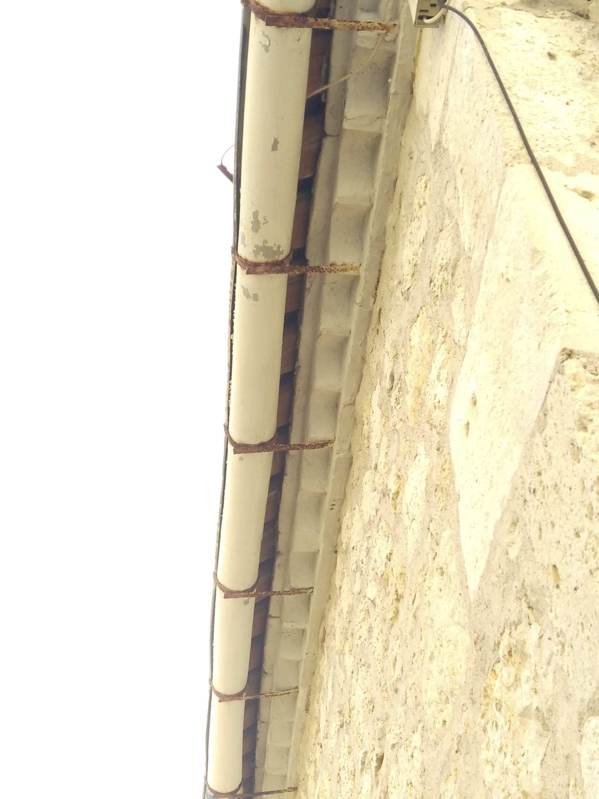 Gouttière crème rouillée avec supports métalliques sur un mur ancien en pierre calcaire, sous un ciel lumineux.