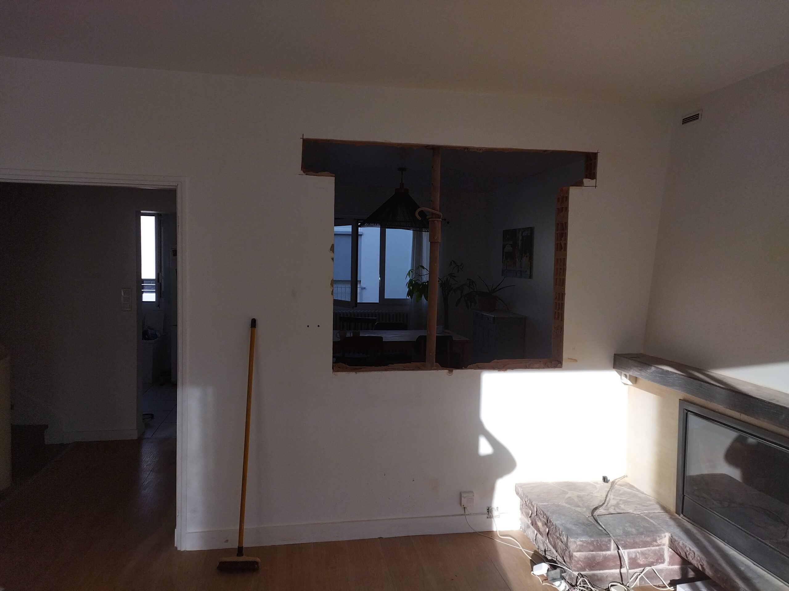 Intérieur de maison en rénovation avec une grande ouverture dans un mur blanc. Vue sur salle à manger, cheminée en pierre et balai.