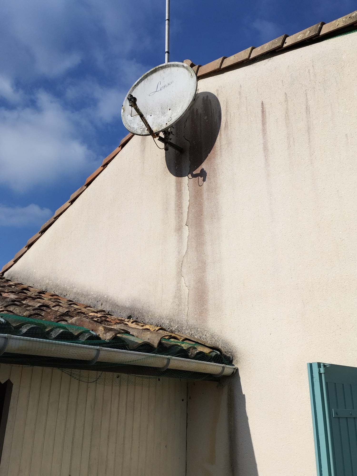 Parabole Lenson sale sur façade beige fissurée et tachée. Toit en tuiles avec filet, gouttière et ciel bleu. Ancien équipement.