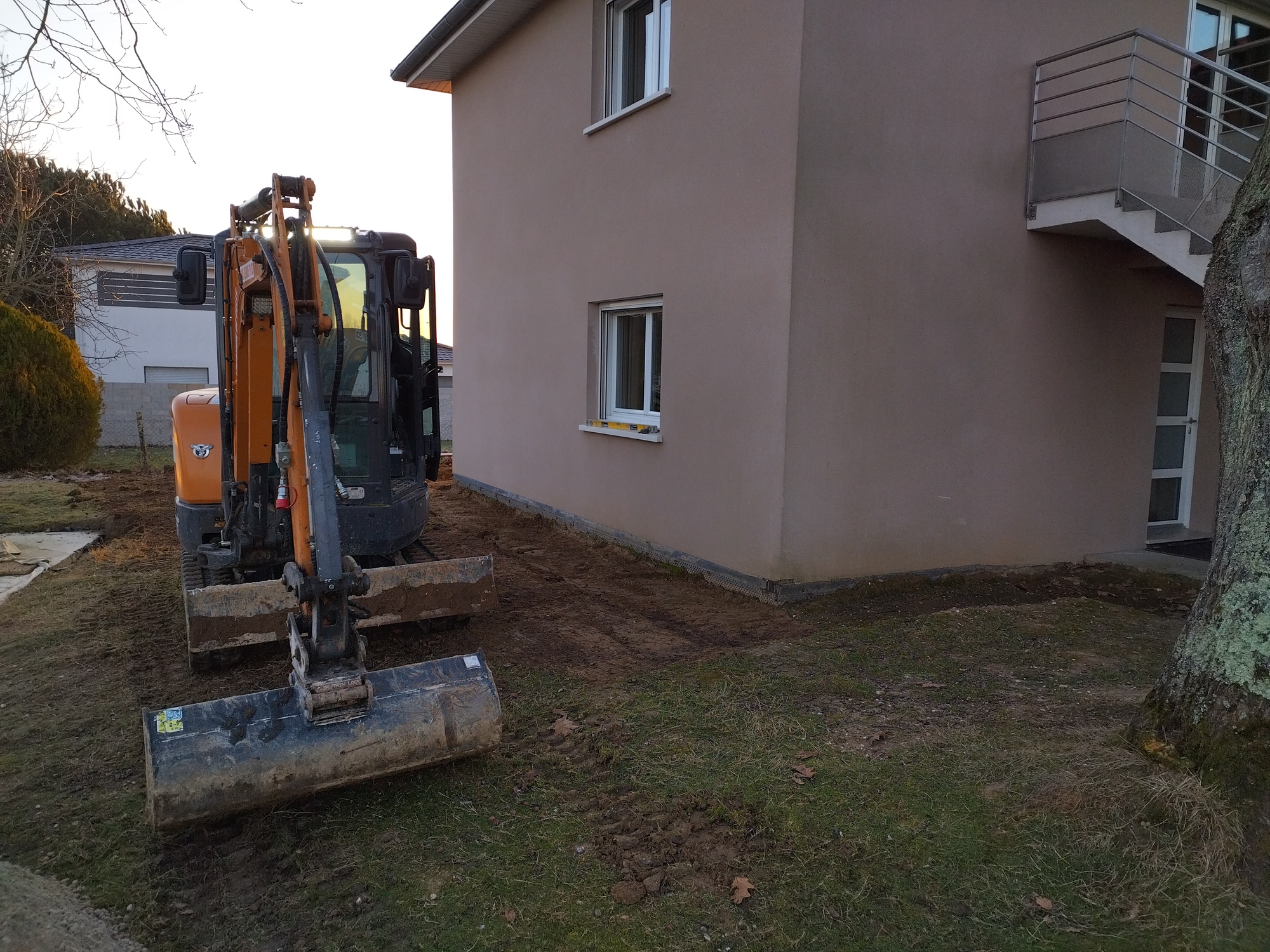 Mini-pelle orange sur terrain déblayé à côté d'une maison moderne beige avec escalier extérieur. Préparation du sol.