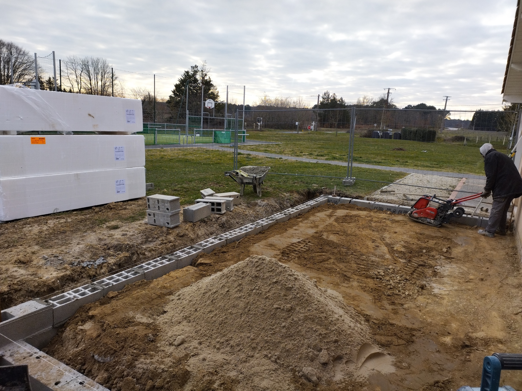 Chantier de fondations avec un homme compactant le sol terreux. Des parpaings et une pile de sable sont visibles, ainsi que des blocs 'RÉSERVÉ'.