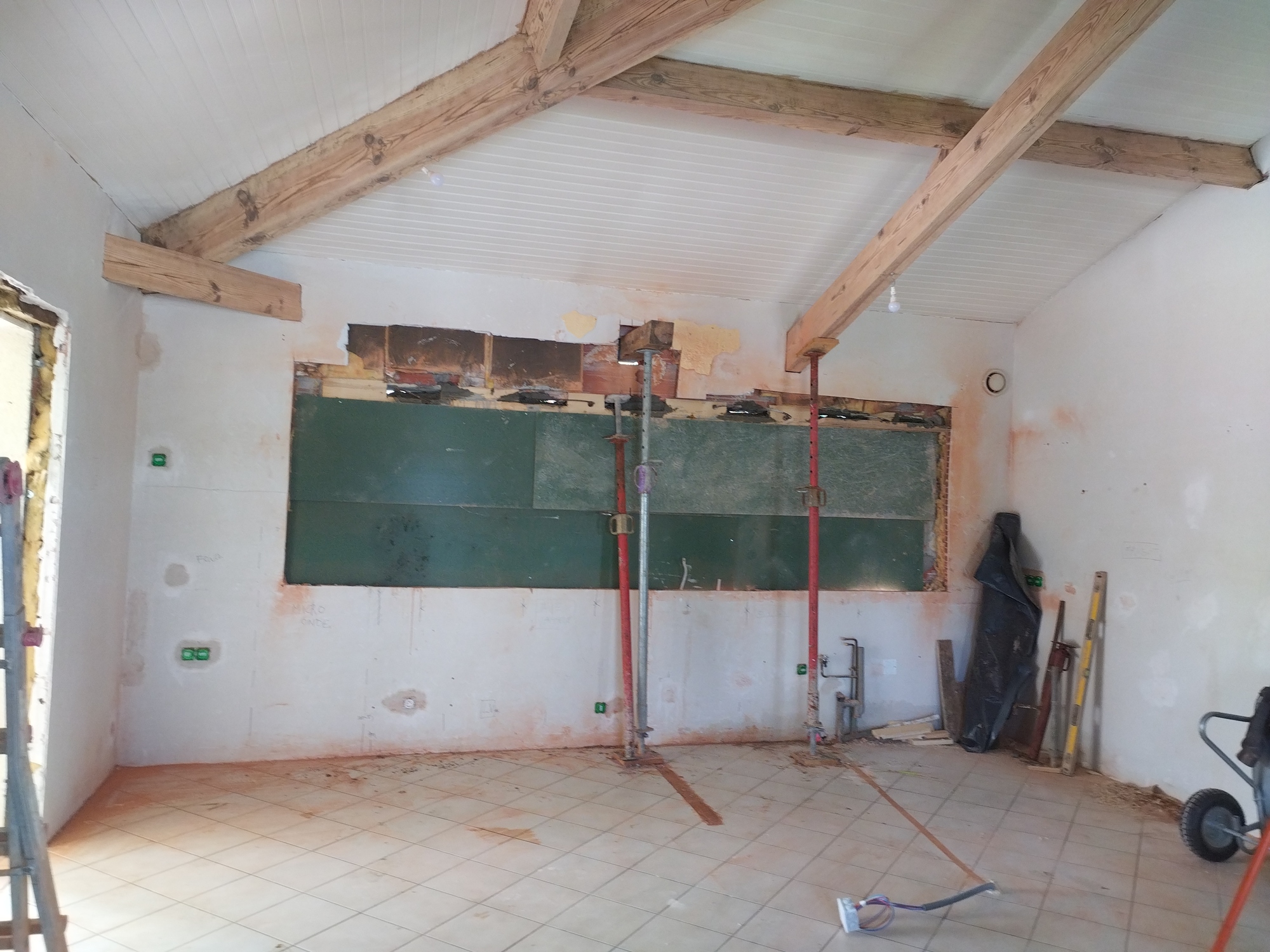 Intérieur en rénovation avec poutres apparentes et plafond lambrissé. Murs en travaux, étais de maçon, carrelage. Texte mural: FUIR, MICRO ONDE.