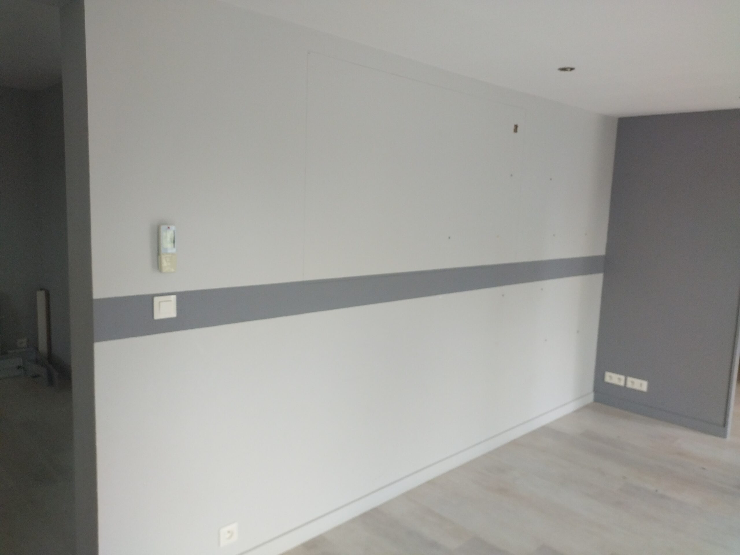 Pièce moderne fraîchement peinte avec murs gris clair et bande horizontale gris foncé. Sol clair, interrupteur mural et prises électriques visibles.