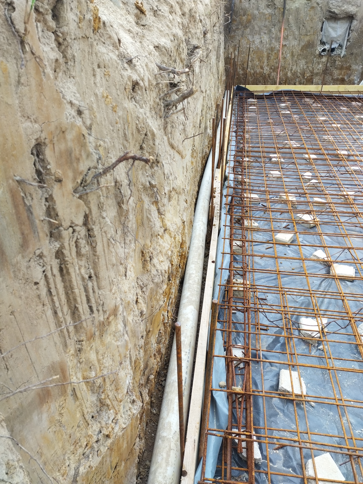 Chantier: tranchée profonde avec mur de terre et tuyau PVC. Armature métallique rouillée posée sur bâche noire, préparant une dalle de fondation.