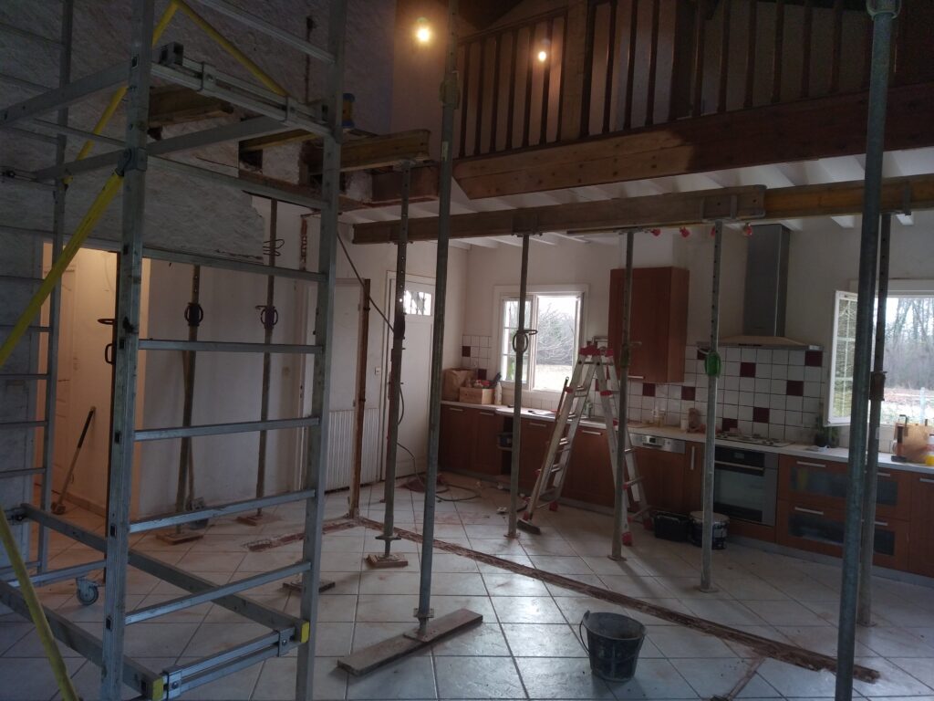 Intérieur de maison en pleine rénovation avec étaiement, échafaudage, cuisine équipée, et mezzanine sous des poutres soutenues.