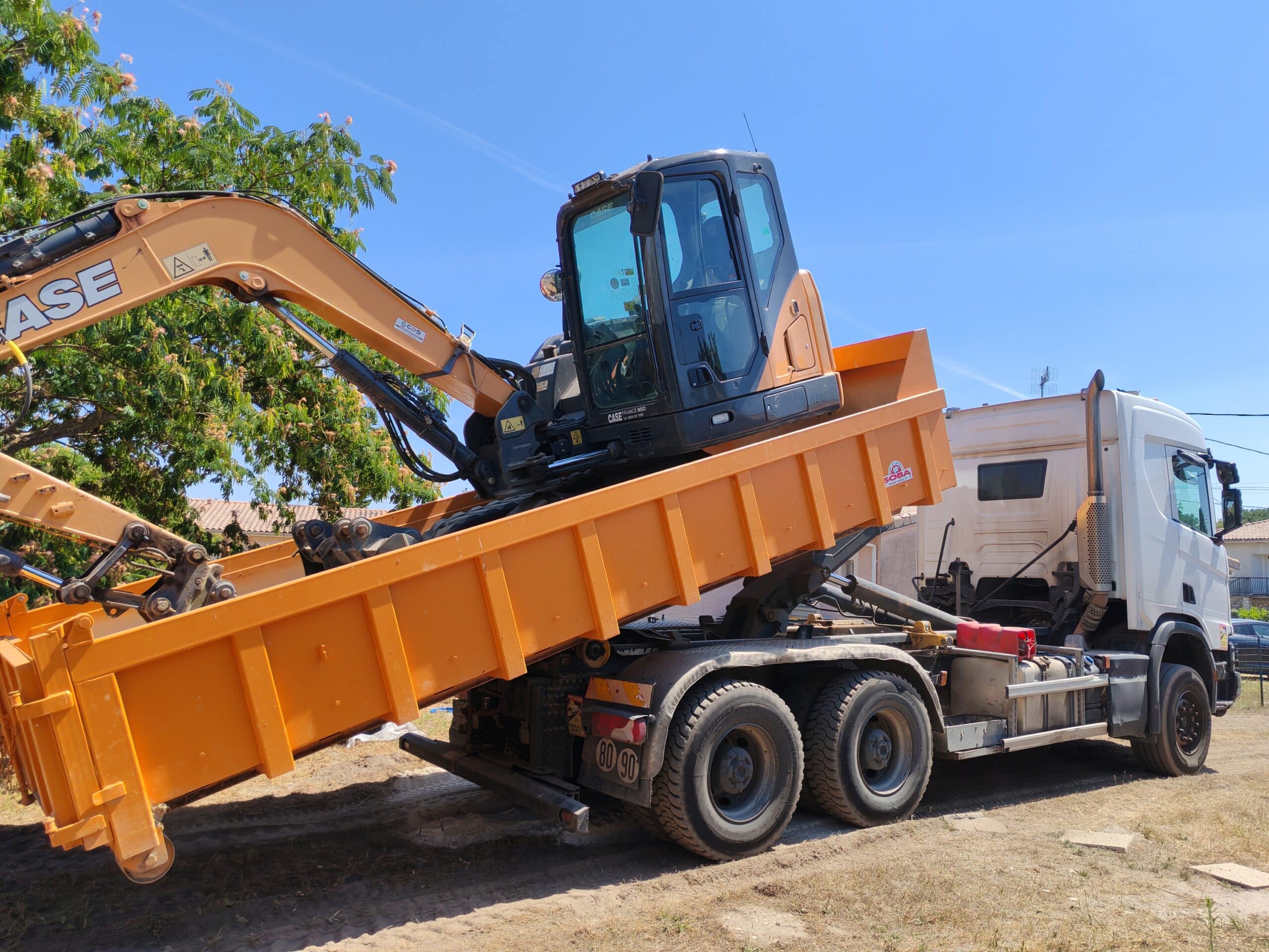 Excavatrice CASE orange sur benne de camion blanche, chargée pour transport. Scène de chantier ensoleillée avec arbres et ciel bleu.