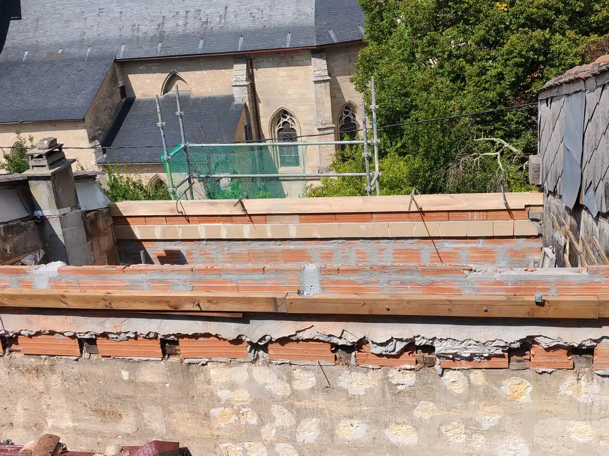 Rénovation de toiture : nouvelles briques et poutres en bois sur un chantier. Vieille église en pierre, toits en ardoise et échafaudage en fond.