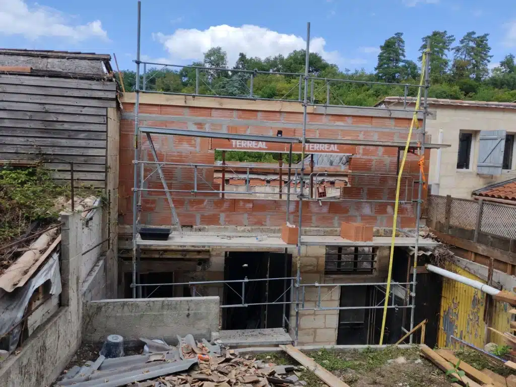 Mur en briques TERREAL en construction avec échafaudage. Vieux bâtiment bois, maison aux volets bleus. Débris de chantier.