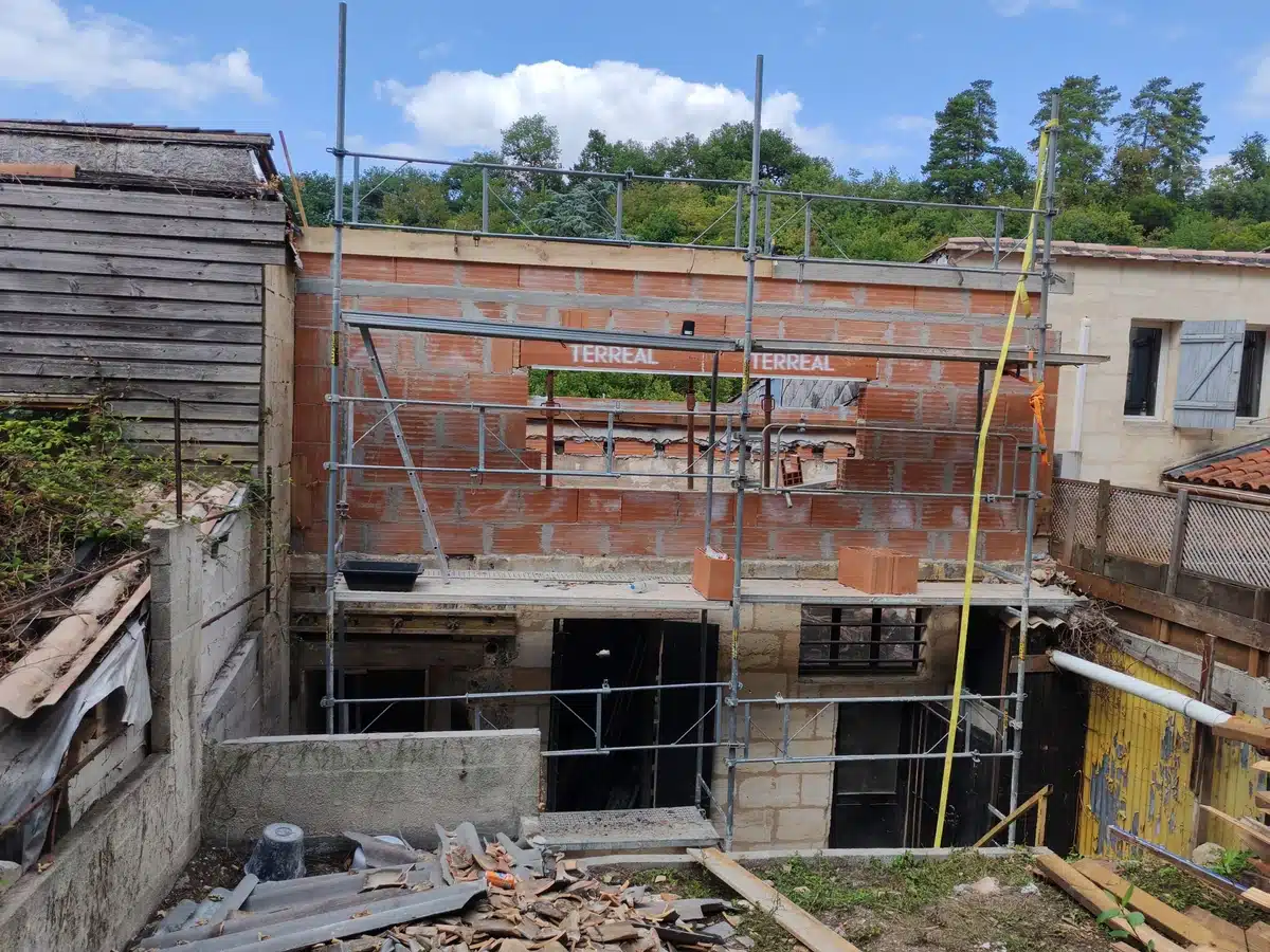 Mur en briques TERREAL en construction avec échafaudage. Vieux bâtiment bois, maison aux volets bleus. Débris de chantier.