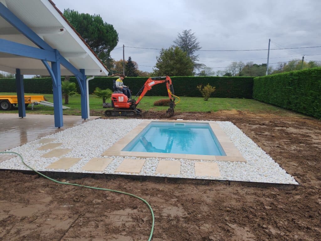 Chantier piscine : mini-pelle Kubota U15-3 près d'un bassin avec margelles beige et graviers blancs. Un ouvrier travaille sur un sol boueux.