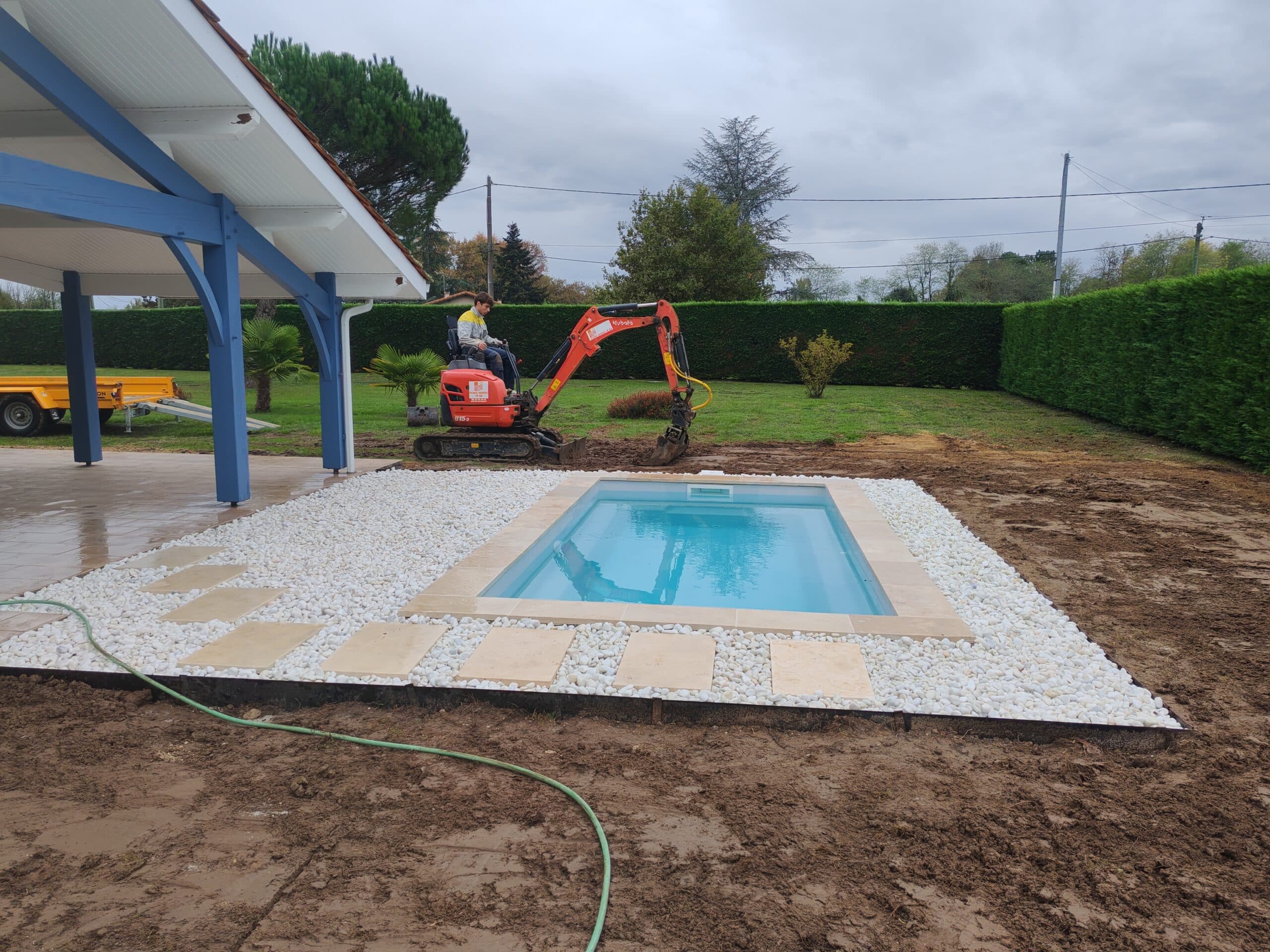 Chantier piscine : mini-pelle Kubota U15-3 près d'un bassin avec margelles beige et graviers blancs. Un ouvrier travaille sur un sol boueux.