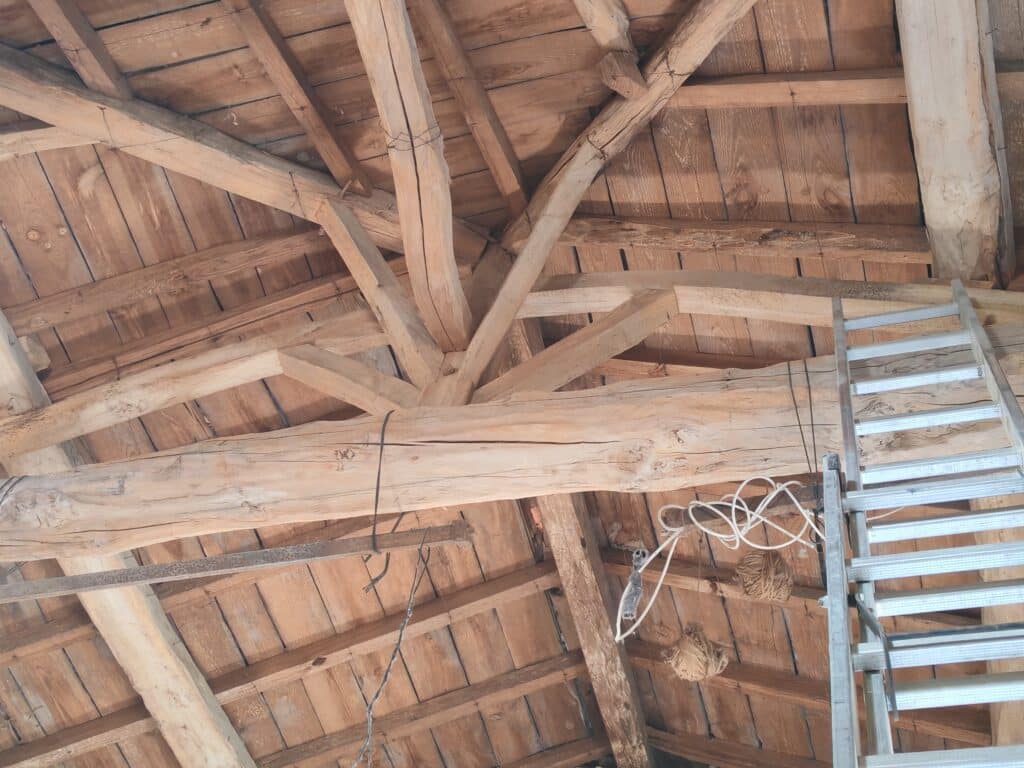 Charpente ancienne en bois clair avec poutres rustiques, escalier métallique et fils électriques pendant, vue de dessous.