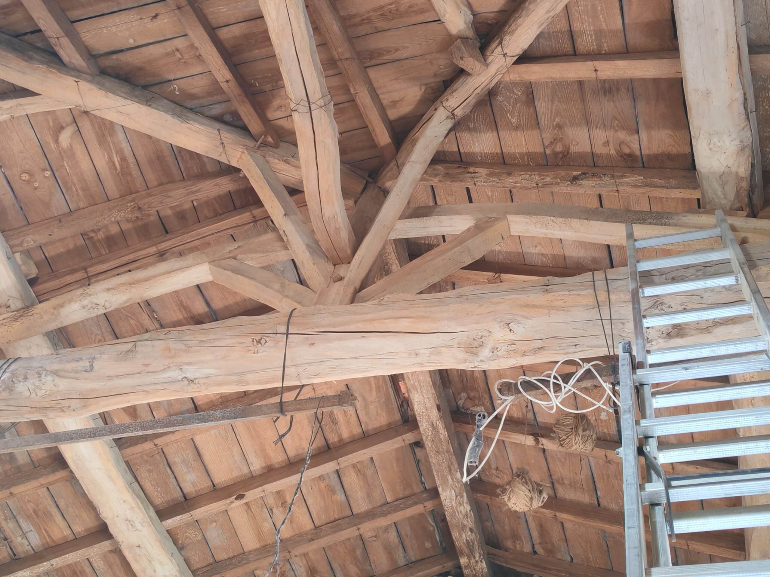 Charpente ancienne en bois clair avec poutres rustiques, escalier métallique et fils électriques pendant, vue de dessous.