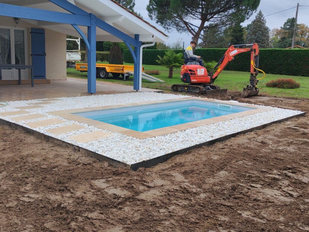 Jeune homme sur mini-pelle Kubota installant une petite piscine avec margelles claires et galets blancs, chantier boueux.
