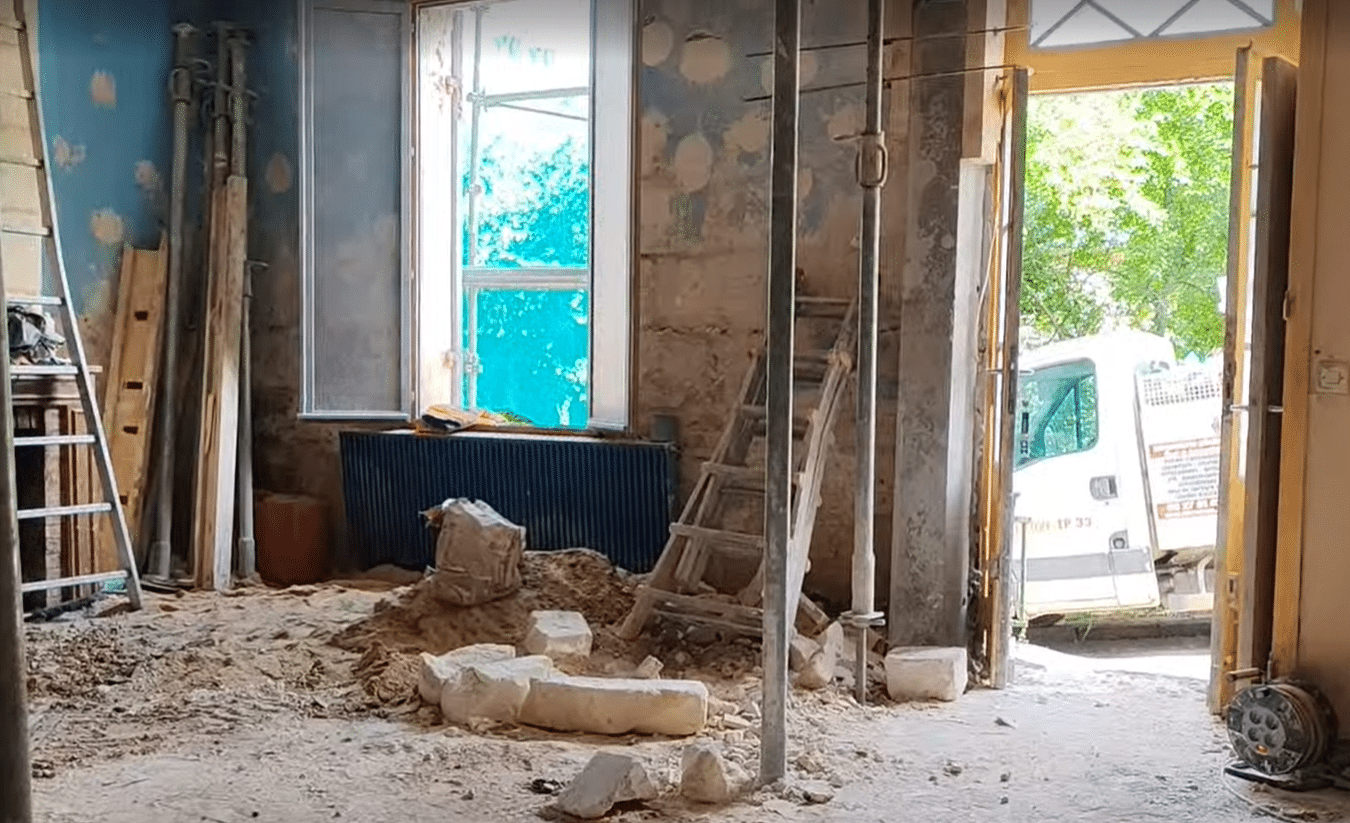 Chantier de rénovation intérieure avec gravats, échelle et étais. Fenêtre et camionnette blanche à l'extérieur.