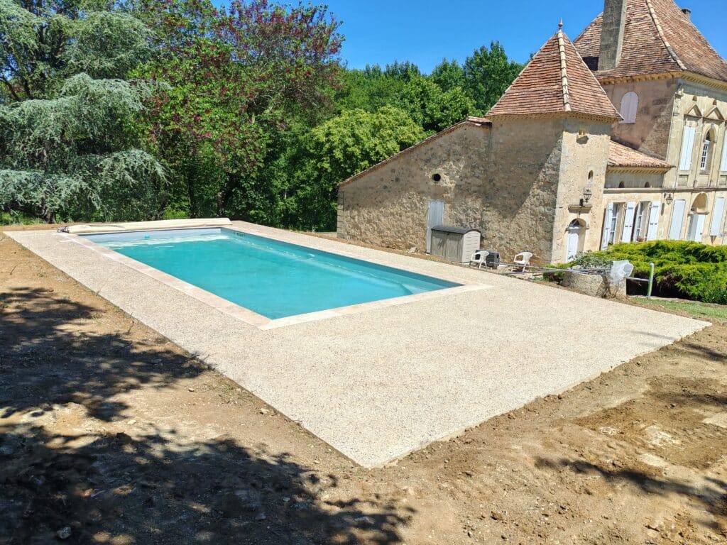 Piscine rectangulaire avec plage en gravier devant un manoir en pierre ancienne sous un ciel bleu d'été.