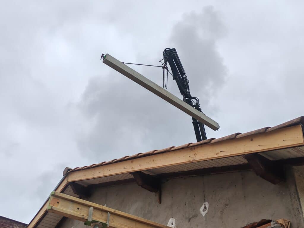 Grue articulée soulevant une poutre sur une toiture en construction.