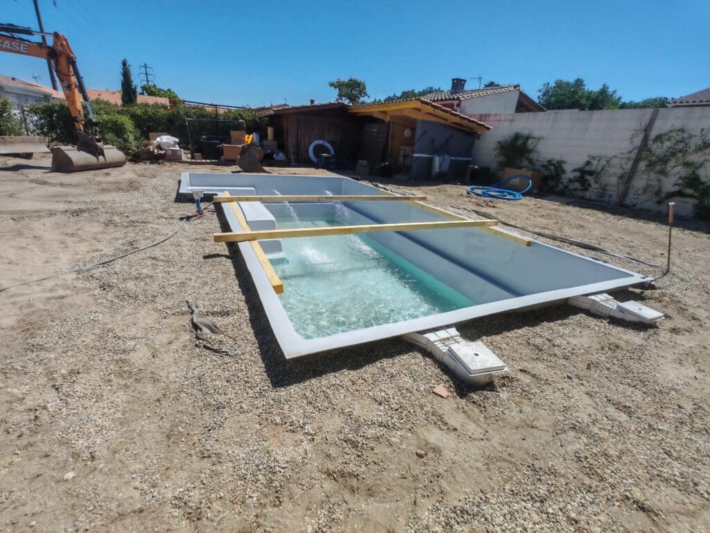 Installation d'une piscine monocoque grise avec pelleteuse Case sur un terrain en gravier sous un ciel bleu.