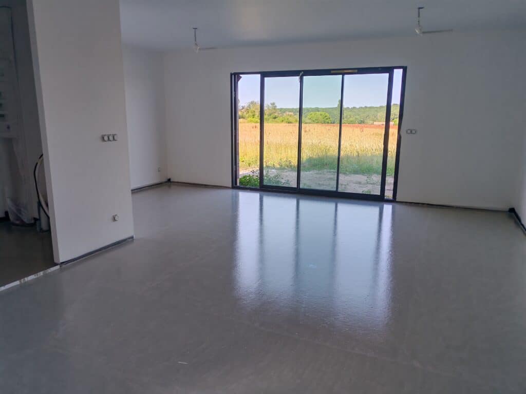 Salon moderne vide avec sol en béton poli et baie vitrée ouvrant sur la nature.